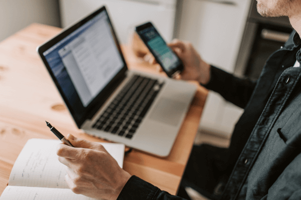 close up of person sitting with a laptop and note pad on the table and a smart phone in the other hand
